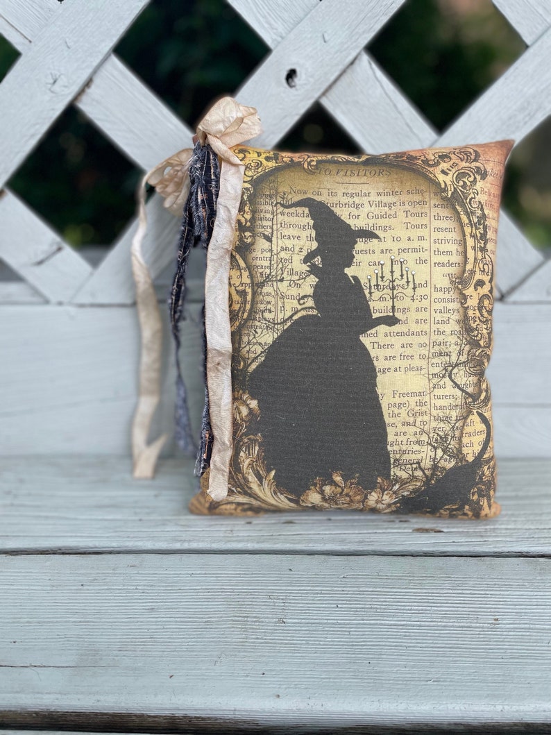 May include: A decorative pillow featuring a silhouette of a witch in a hat and dress, set against a vintage map background. The pillow has a cream-colored ribbon and fabric accents. The text on the pillow reads "TO VISITORS".