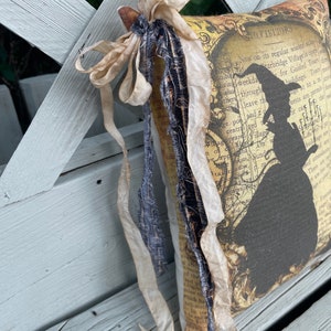 May include: A decorative pillow with a vintage aesthetic, featuring a silhouette of a witch and text. The pillow is adorned with cream-colored ribbons and a dark blue fabric accent. The background appears to be a weathered wooden bench.
