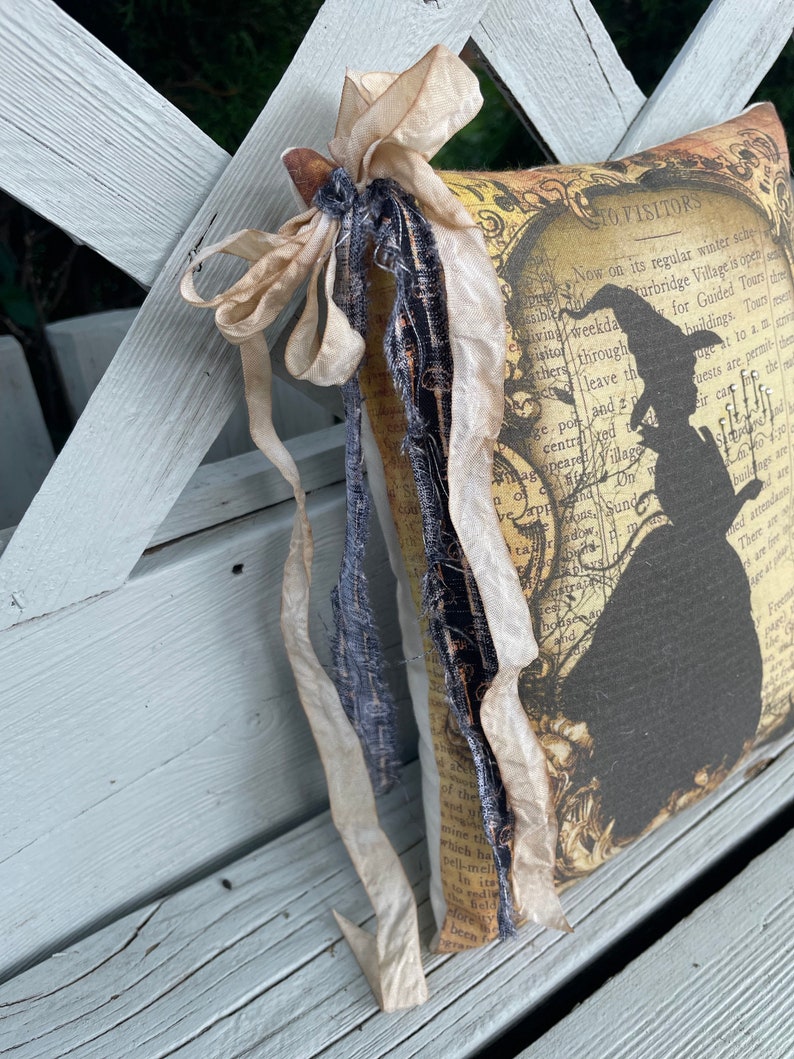 May include: A decorative pillow with a vintage aesthetic, featuring a silhouette of a witch and text. The pillow is adorned with cream-colored ribbons and a dark blue fabric accent. The background appears to be a weathered wooden bench.