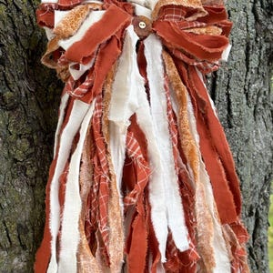 May include: A decorative hanging ornament crafted from numerous strips of fabric in shades of burnt orange, cream, and white. The fabric pieces feature solid colors, plaid patterns, and floral prints, all gathered at the top with a small wooden button.