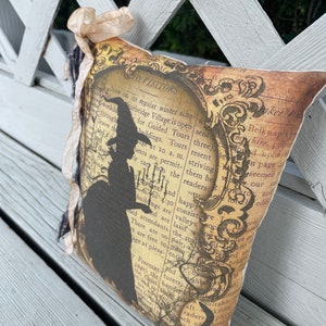 May include: Decorative pillow with a vintage-style design. The pillow features a silhouette of a witch and text on a background resembling aged paper. The pillow is embellished with a beige ribbon and dark fringe.