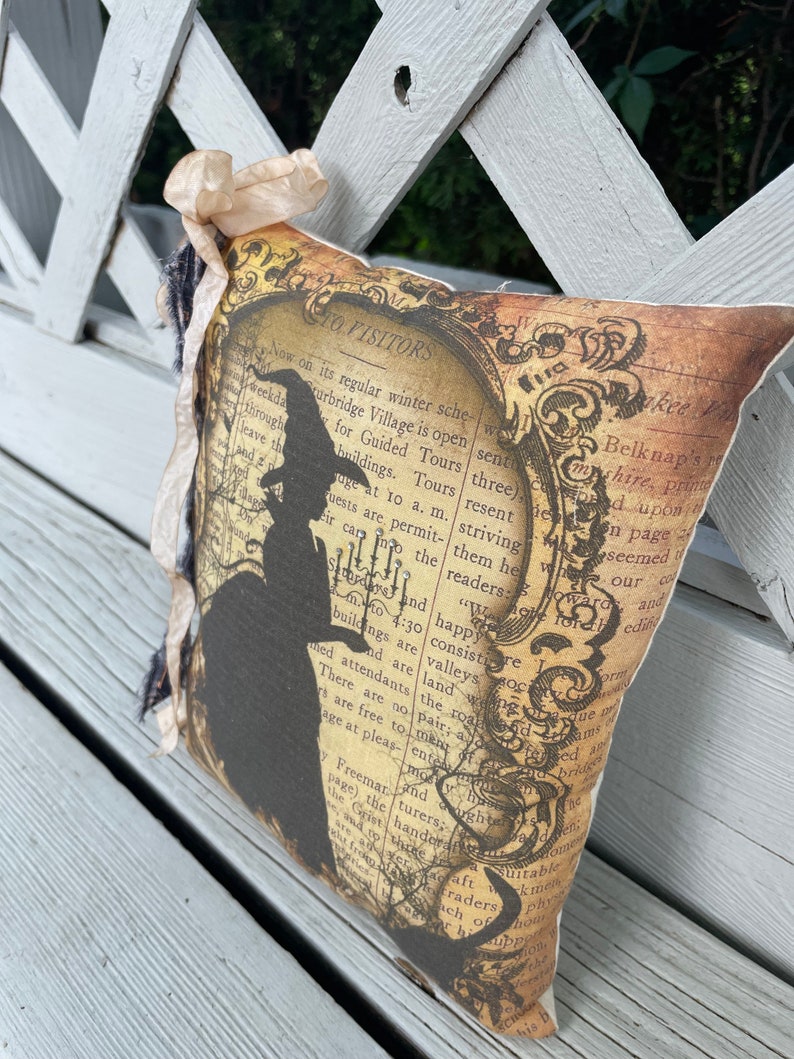 May include: Decorative pillow with a vintage-style design. The pillow features a silhouette of a witch and text on a background resembling aged paper. The pillow is embellished with a beige ribbon and dark fringe.