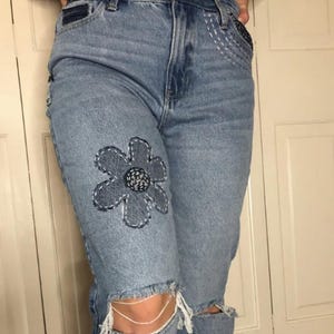 May include: Light blue denim jeans with a large, dark blue embroidered flower on the left knee. The jeans have a distressed look with rips and tears.