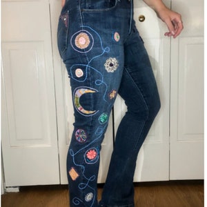May include: Blue denim jeans with embroidered embellishments, including a crescent moon, stars, and flowers.