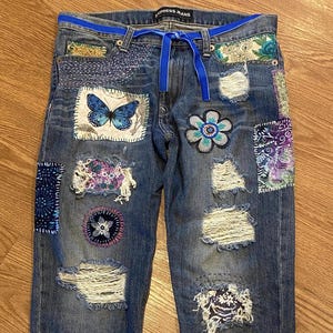 May include: Blue denim jeans with various patches and embroidery. The patches include a butterfly, a flower, and a paisley design. The jeans have a blue ribbon tied at the waist.