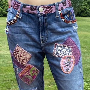 May include: Blue denim jeans with various fabric patches in shades of purple, brown, and green. The jeans have a pink and brown patterned belt.