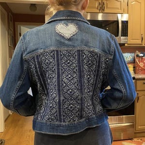 May include: A blue denim jacket with a white heart patch on the back. The back of the jacket features a decorative pattern of blue and white embroidery.