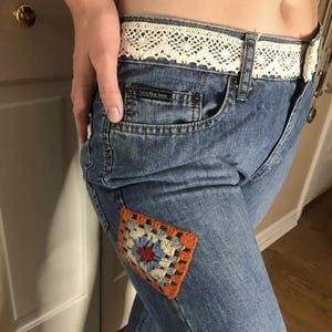 May include: Blue denim jeans with a crocheted patch on the left leg. The jeans have a white lace waistband and a Calvin Klein Jeans label on the back pocket.