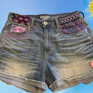 May include: Denizen High Rise denim shorts with embroidered details in pink, purple, and blue. The shorts have a frayed hem and a pink flower patch.