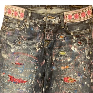 May include: Dark wash denim jeans with a decorative waistband featuring a pink floral pattern. The jeans have colorful paint splatters and visible stitching repairs. The text "LIL MAGGIE" is visible on the waistband.