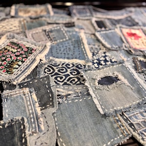 May include: A collection of denim patches in various shades of blue, arranged closely together. Some patches feature floral and patterned fabric inserts, while others have visible white stitching and frayed edges. The overall effect is a textured, handcrafted appearance.