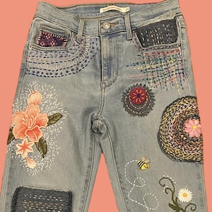 May include: Light blue denim jeans with colorful embroidered floral and geometric designs. Features include a large pink flower, a bee, and stitched patches. The jeans have a button and zipper closure.