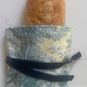 May include: A green and white patterned fabric bread bag with a black drawstring. The bag is filled with a loaf of bread.