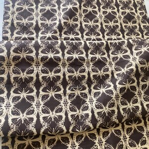 May include: Close-up of a fabric with a repeating butterfly pattern. The butterflies are cream-colored and outlined on a dark brown background. The fabric is folded over, showing the pattern in multiple layers.