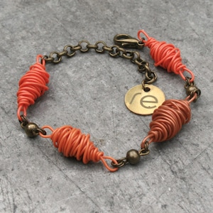 Pode incluir: Um bracelete de fio enrolado cor laranja coral com uma corrente de latão e um amuleto de latão com a letra "re" nele.