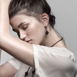May include: A woman with dark brown hair wearing a white t-shirt and a black choker necklace with a pendant. She is wearing a pair of black earrings.