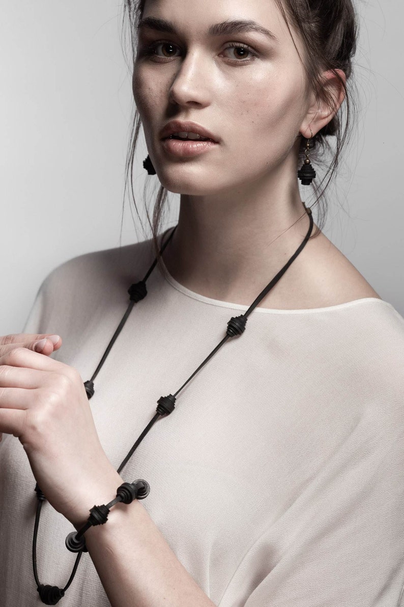 May include: A woman wearing a white top with a black necklace and bracelet. The necklace and bracelet are made of layered black circles.