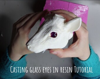 May include: A white resin deer head with purple eyes is held in hands. The head is textured and detailed, with visible ears and facial features. The text "CASTING GLASS EYES IN RESIN TUTORIAL" is at the bottom of the image.
