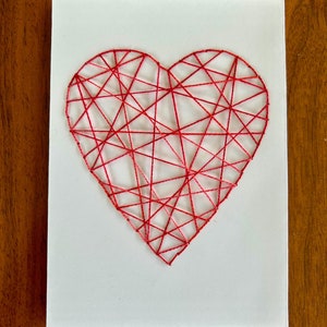 May include: A white card with a red string heart design. The heart is made of string wrapped around nails in a geometric pattern.