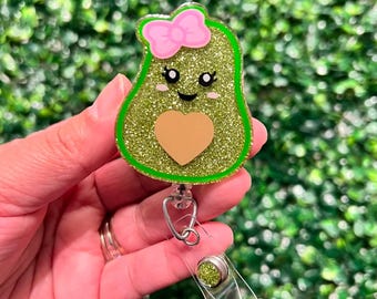 May include: A green glitter avocado with a pink bow and a gold glitter heart, attached to a clear plastic badge holder with a silver clip.