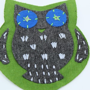May include: A handmade owl applique with a gray body, blue eyes, and white accents, set on a green background. Includes a small group of bronze-colored metal studs. Ideal for crafting or embellishing projects.