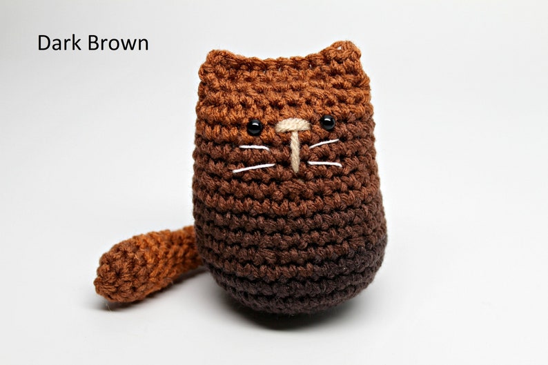 May include: A handmade crochet cat in shades of dark brown. The cat has black eyes, white whiskers, and a beige nose. The text "Dark Brown" is in the upper left corner. The cat has a small tail.