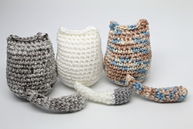 May include: Three handmade crochet cat toys in gray, white, and a mix of blue and brown. Each cat toy has a tail and a rounded body with pointed ears. The toys are displayed on a white background.