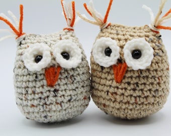 May include: Two crocheted owls, one white with orange accents and one brown with orange accents. Both owls have white eyes with black pupils.