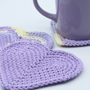 May include: Three crocheted heart-shaped coasters in shades of purple and yellow. The coasters are made with a textured stitch and are perfect for protecting surfaces from hot drinks.