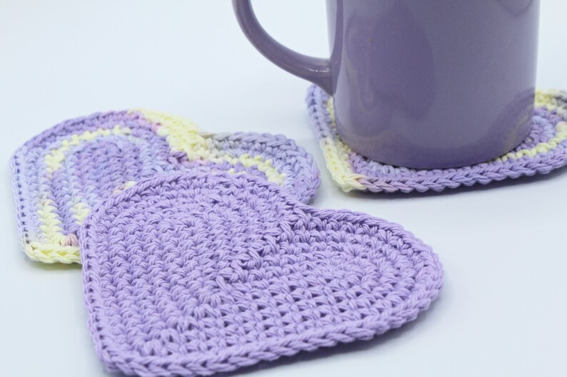 May include: Three crocheted heart-shaped coasters in shades of purple and yellow. The coasters are made with a textured stitch and are perfect for protecting surfaces from hot drinks.