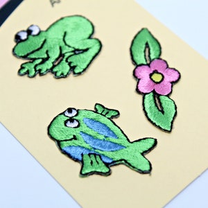 以下が含まれることがあります： 緑色のカエル、青い縞模様の魚、緑の葉とピンクの花の刺繍ワッペン。ワッペンは「Next Style」のロゴが入った薄い黄色のカードにあります。
