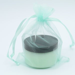 May include: A light green tulle bag with a drawstring closure, containing a white jar with a black lid. The jar is filled with a white cream.