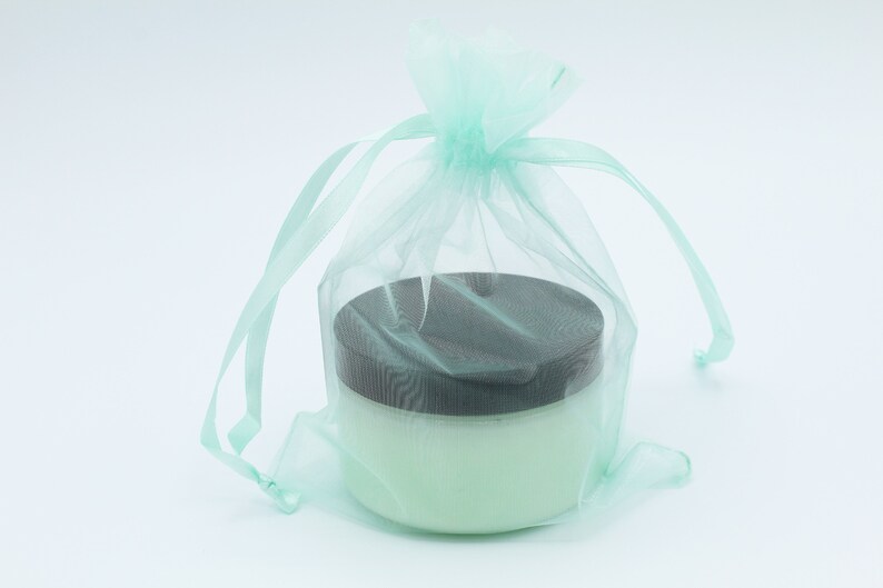 May include: A light green tulle bag with a drawstring closure, containing a white jar with a black lid. The jar is filled with a white cream.