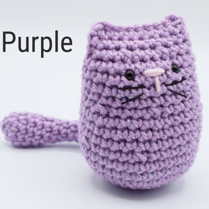 May include: Purple crocheted cat with black eyes, pink nose, and black whiskers. The cat is sitting on a white surface.