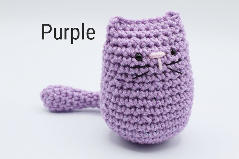 May include: Purple crocheted cat with black eyes, pink nose, and black whiskers. The cat is sitting on a white surface.