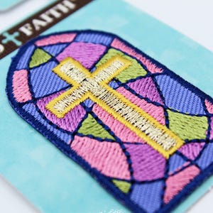 May include: Embroidered patch with a stained-glass window design and a gold cross. The patch has a blue border and is filled with pink, purple, green, and blue sections. The words "BOLD + FAITH" are printed on the packaging.