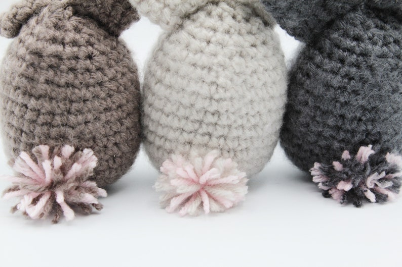 May include: Three crocheted stuffed animals with pink pom-pom tails. The animals are brown, gray, and dark gray.