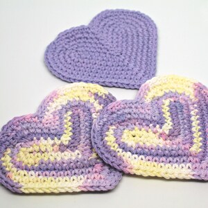 May include: Three crocheted heart-shaped coasters in shades of purple, pink, and yellow. The coasters are made with a textured stitch and have a slightly raised edge.