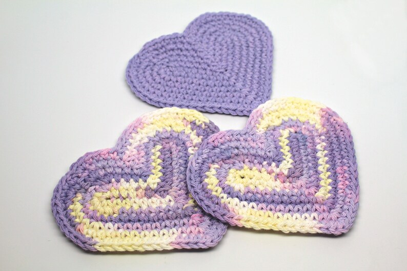 May include: Three crocheted heart-shaped coasters in shades of purple, pink, and yellow. The coasters are made with a textured stitch and have a slightly raised edge.