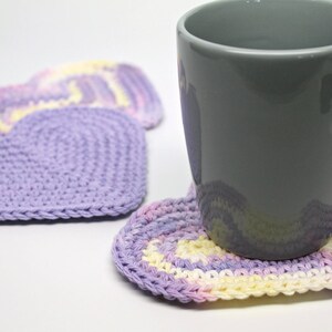 May include: A gray ceramic mug resting on a purple and yellow crocheted heart-shaped coaster. Two additional heart-shaped coasters are visible in the background.