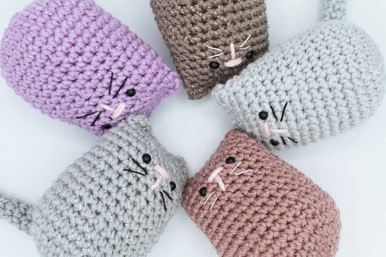 May include: Five crocheted cat toys in different colors, including purple, brown, gray, and pink. Each cat toy has a black embroidered face with pink nose and whiskers.
