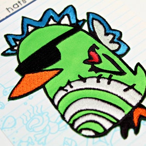 Op de afbeelding: Groen, blauw en wit geborduurd patch van een cartoonmonster met een zwart ooglapje en een oranje snavel. Het monster heeft een gestikt lichaam en een rood hart op zijn borst.