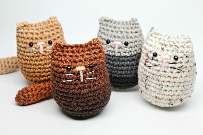 May include: Four handmade crochet cat toys in shades of brown, gray, and speckled white. Each cat has black eyes, a pink nose, and white whiskers. The cats are standing upright, with one cat having a tail.