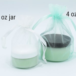 May include: Two jars of green cream in white and light green organza bags. The smaller jar is labelled "2 oz jar" and the larger jar is labelled "4 oz jar".