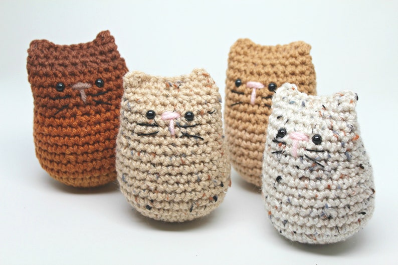 May include: Four handmade crochet cat figures in various colors: brown, tan, and speckled white. Each cat has black eyes, a pink nose, and black whiskers. The figures are soft and rounded, perfect for a child's toy or home decor.