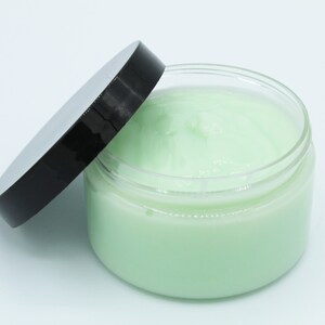 May include: A clear jar with a black lid, partially open, filled with a light green cream.