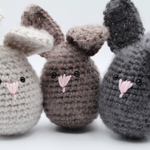 May include: Three crocheted bunny plushies in different colors: light gray, brown, and gray. Each bunny has a pink nose and black eyes.