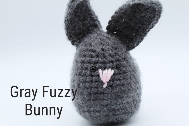 May include: Gray crocheted bunny with pink nose and black eyes. The bunny is sitting on a white surface. "Gray Fuzzy Bunny" is written in black text below the bunny.