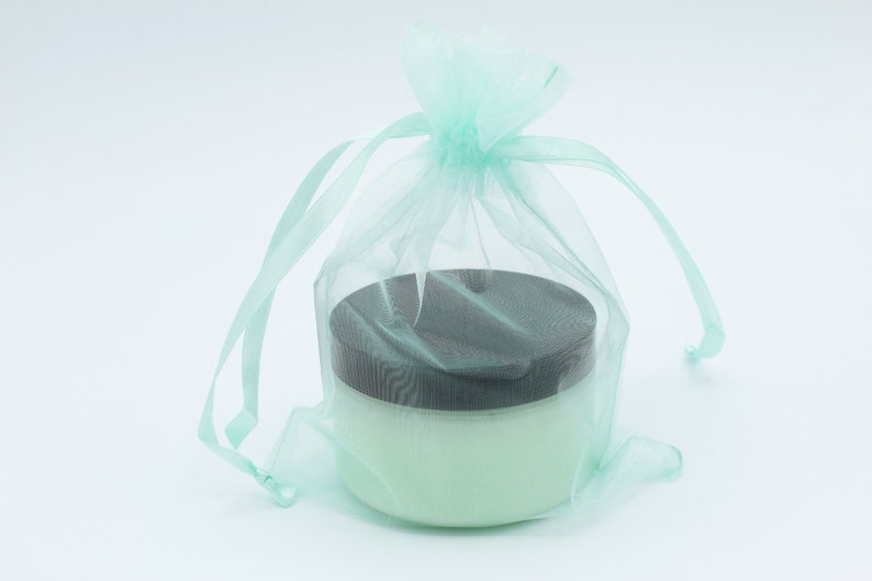 May include: A light green tulle bag with a drawstring closure, containing a white jar with a black lid. The jar is filled with a white cream.