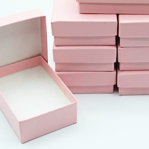 以下が含まれることがあります： ピンクの厚紙製の箱が10個積み重ねられており、1つの箱が開いて横向きになっています。開いた箱の内側は白です。